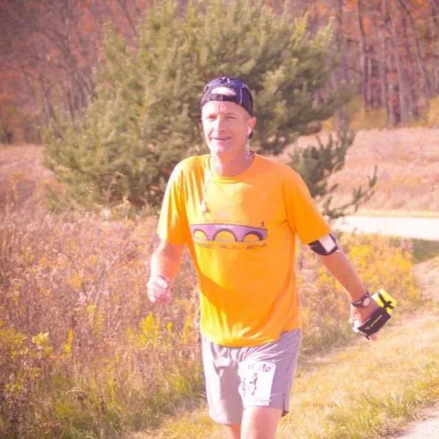 A male runner wearing an orange t-shirt with a bridge graphic and grey shorts runs on a path with trees and dry grass in the background.