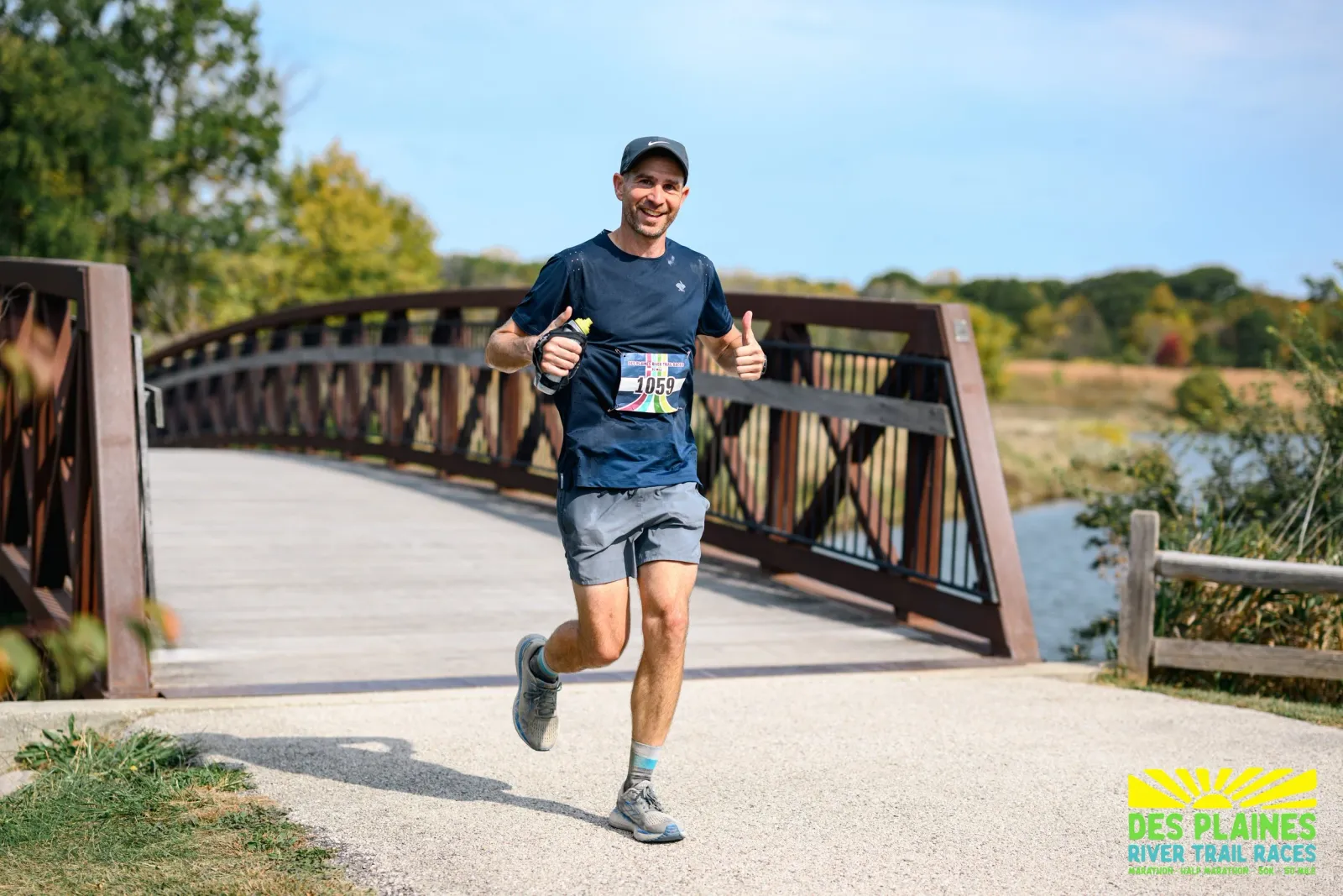 A male runner smiles and gives a thumbs up while running across a wooden bridge on a sunny day.