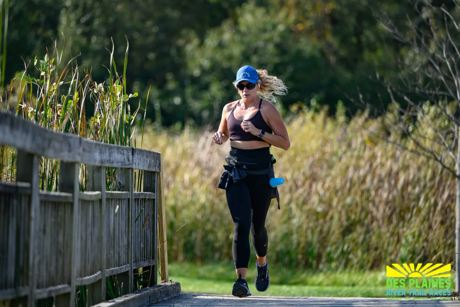 A female runner with blonde hair tied back in a ponytail, wearing a blue cap and sunglasses, runs on a wooden boardwalk next to a wooden railing. She is wearing a dark sports bra and black leggings. The background is filled with tall grasses and trees.