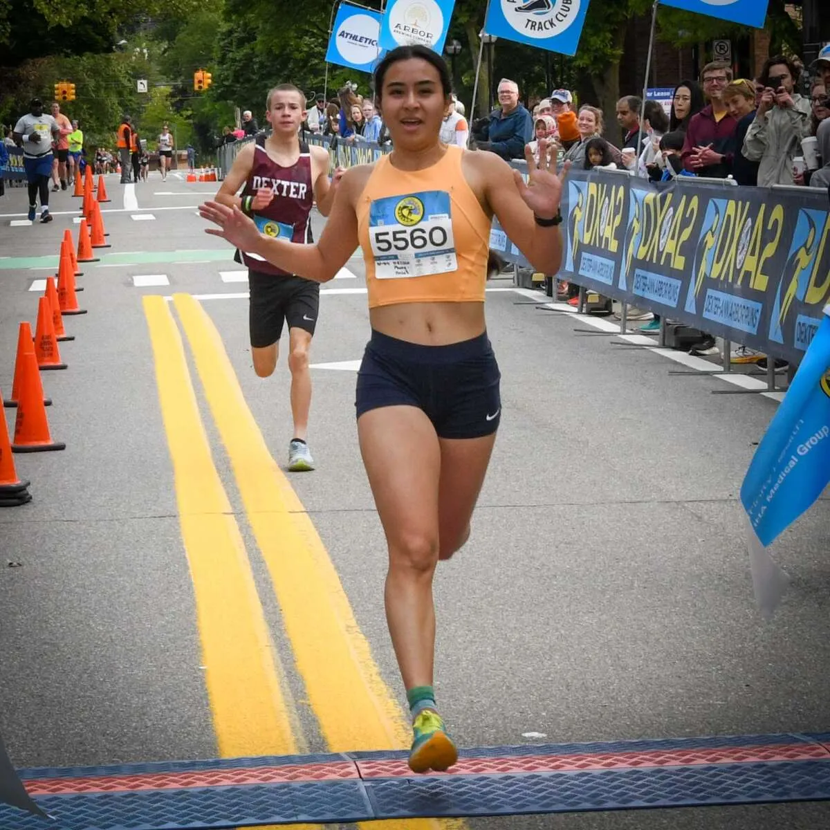 A female runner wearing an orange crop top and dark shorts waves as she crosses the finish line, with another runner behind her and spectators watching.