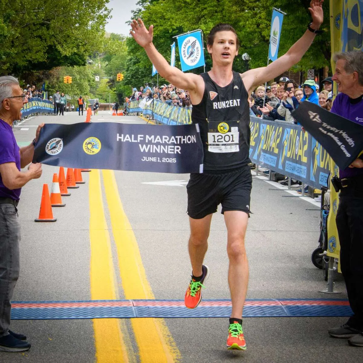 A male runner with his arms raised in victory crosses the finish line of a half marathon, holding a banner that reads 'HALF MARATHON WINNER'.