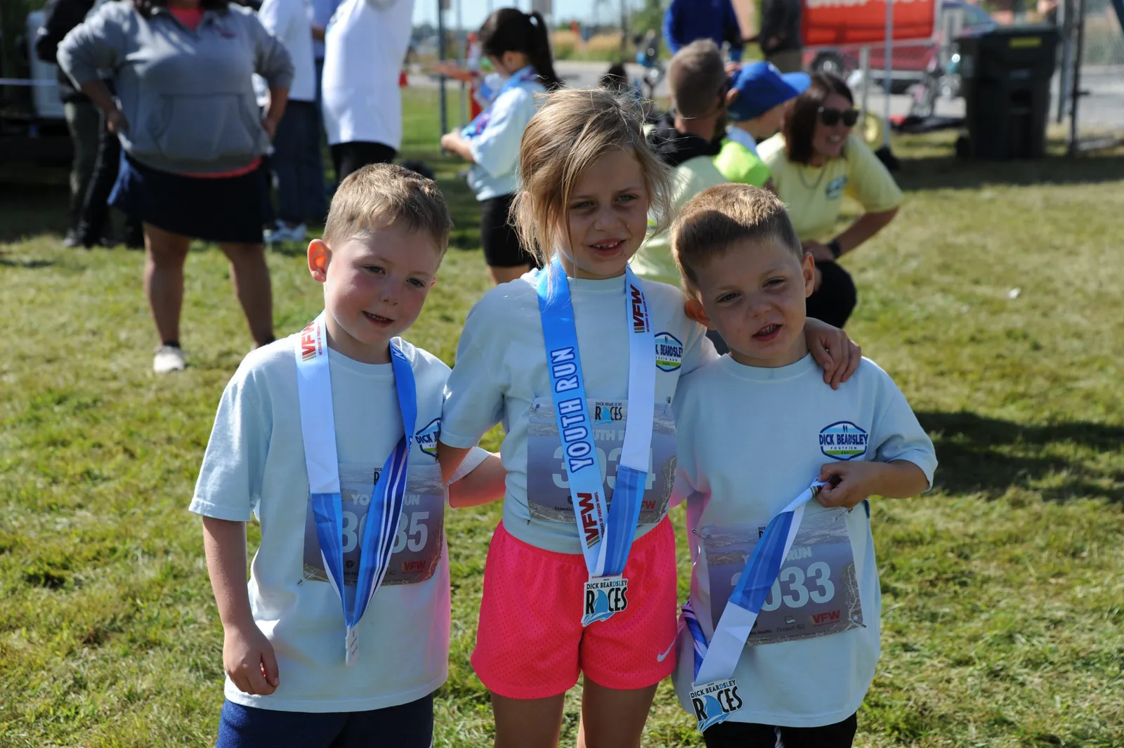 Three young children, two boys and a girl, proudly wearing medals and race bibs from the Dick Beardsley Races Youth Run.