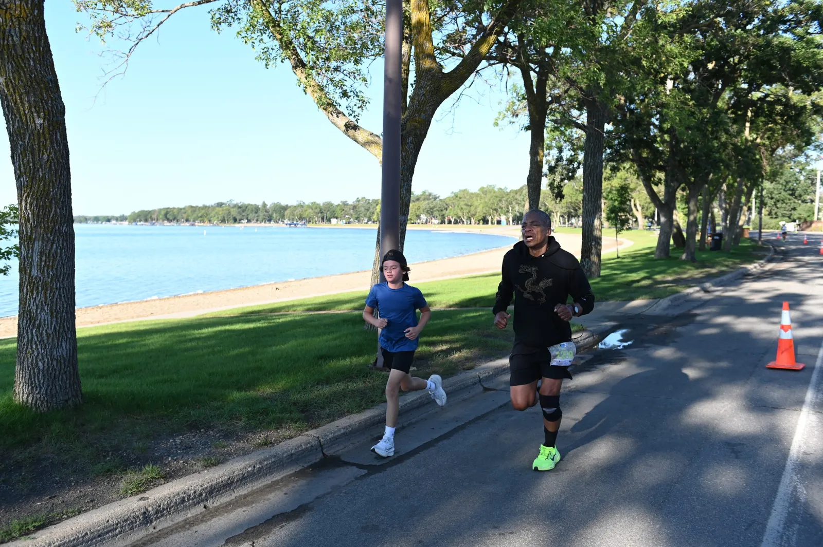 Two runners, one adult and one child, race along a paved path next to a lake.