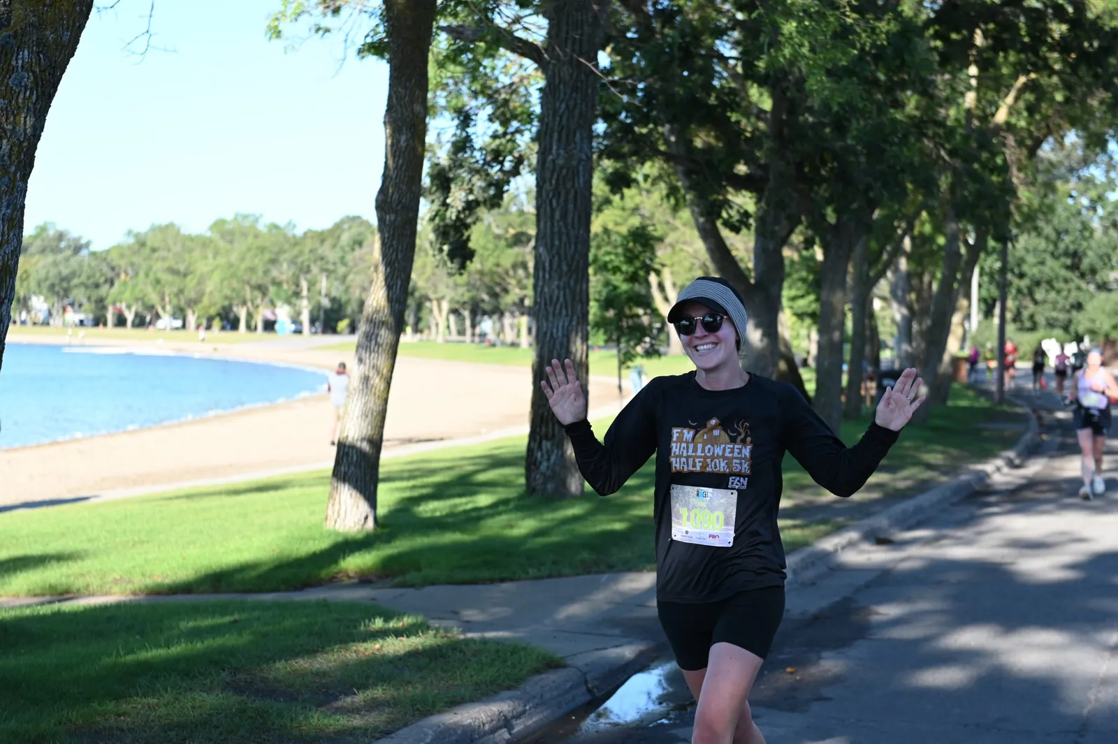 A female runner wearing a black long-sleeved shirt and shorts smiles and waves with both hands while running on a road next to a body of water and a beach.