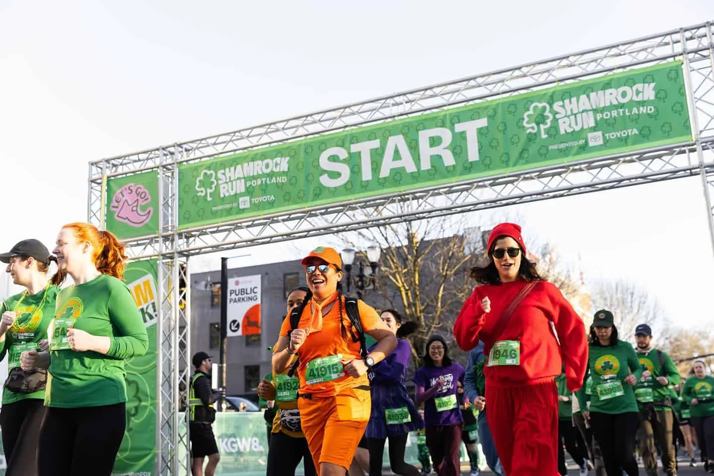 Runners in festive green and red outfits smiling and running under the 'START' banner at the Shamrock Run Portland.