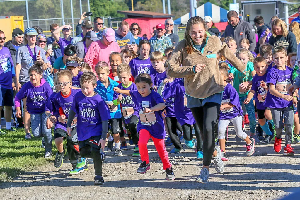 A group of children in purple shirts are running forward at the start of a race, with an adult woman in a tan fleece leading them.