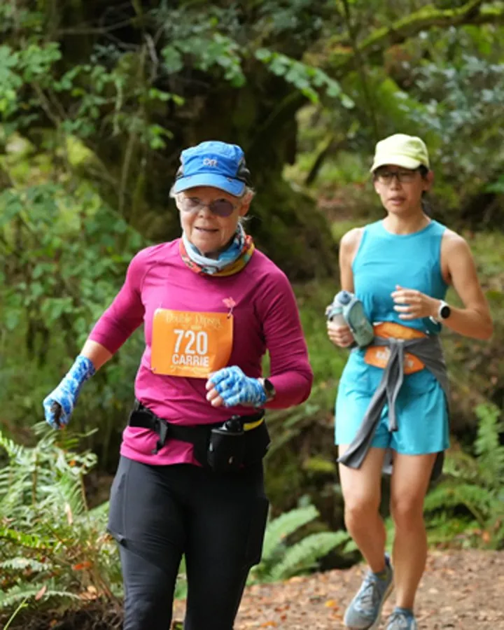 Two female runners on a wooded trail, one in a pink shirt with bib number 720 and the other in a blue tank top.