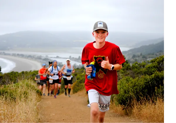 A young runner in a red shirt and a baseball cap smiles and gives a thumbs up while running on a dirt trail, with other runners and a scenic coastal view in the background.