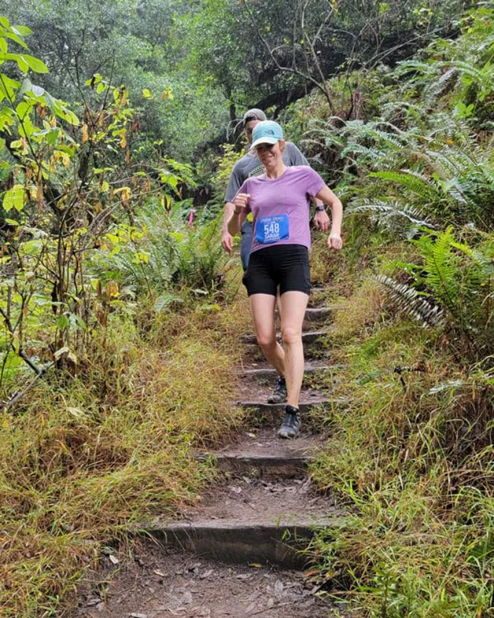 A female runner with bib number 548 descends a wooden staircase on a wooded trail.