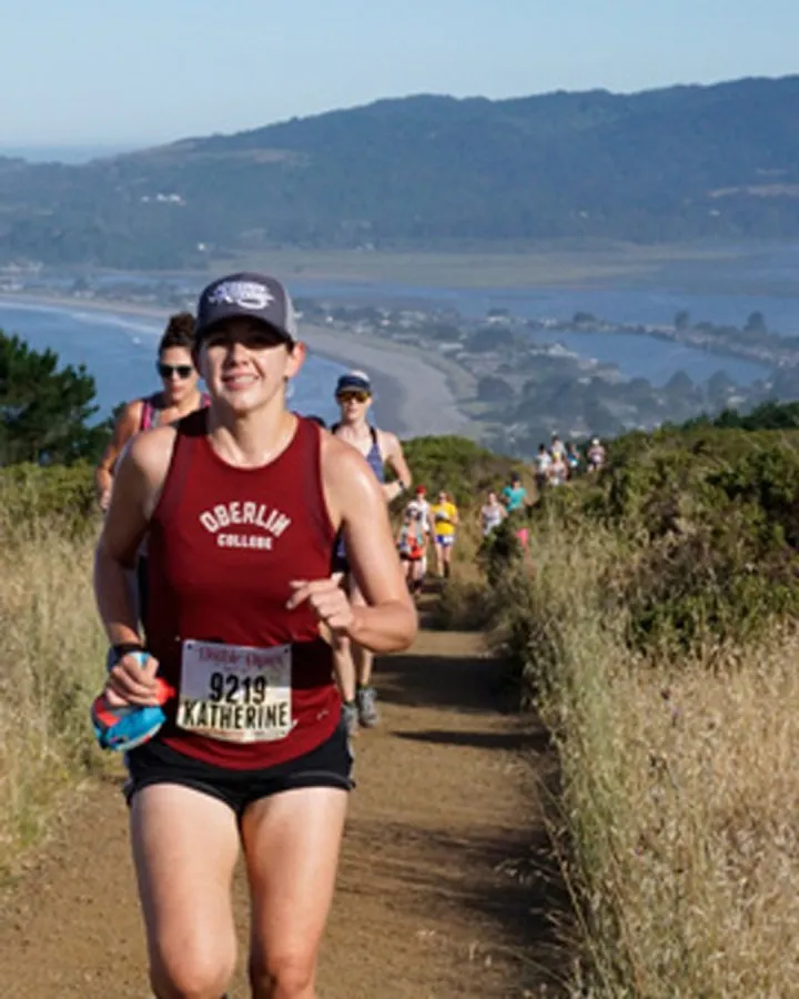 A female runner wearing a maroon Oberlin College singlet and a grey baseball cap runs on a dirt path, with other runners behind her and a scenic coastal view in the background.