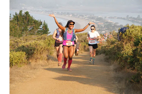 A female runner with arms raised in celebration runs up a dirt trail, with other runners behind her and a scenic coastal view in the background.