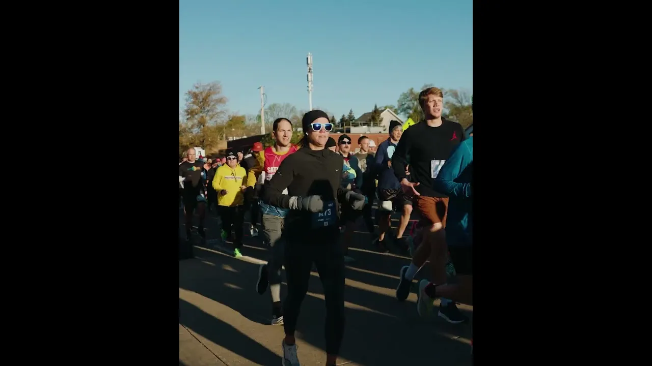 A group of runners starting a race, with a woman in the foreground wearing sunglasses and a black long-sleeved shirt.