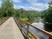 A paved trail bridge crosses a calm river, with lush green trees and rolling hills in the background under a partly cloudy sky.