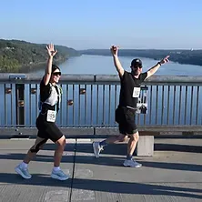 Two runners, a man and a woman, celebrating with their arms raised while running across a bridge with a river and hills in the background.