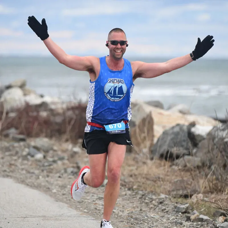 A male runner with arms outstretched in celebration, wearing a blue 'Shipyard' tank top and black shorts, running along a waterfront path.