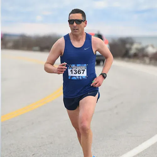 A male runner wearing sunglasses and a blue tank top with bib number 1367 runs on a road with the ocean in the background.