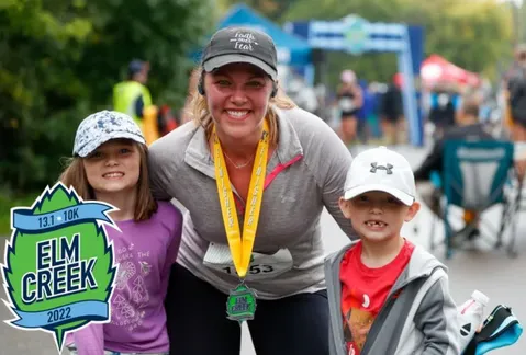 A smiling woman wearing a race medal poses with two children in front of a finish line archway.