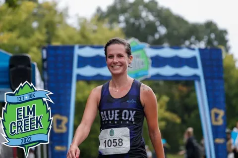 A smiling female runner wearing a blue tank top with 'Blue Ox Running' and bib number 1058 crosses the finish line at the Elm Creek 2022 race.