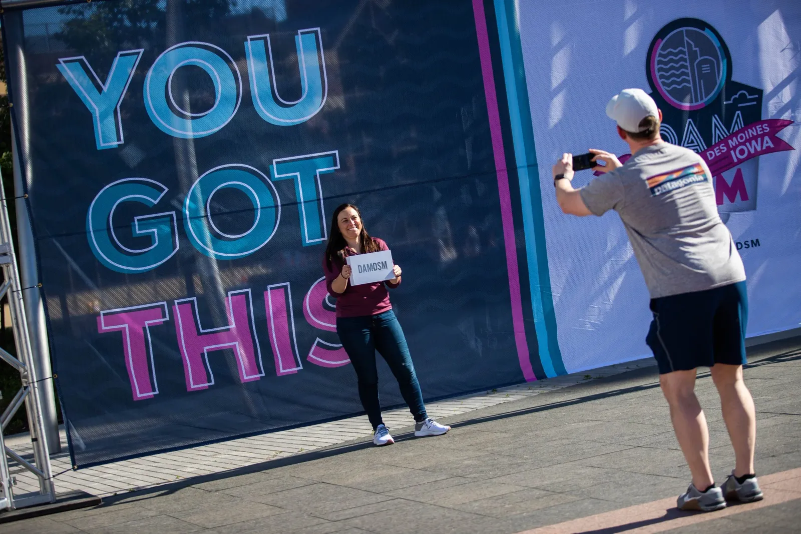 A woman smiles while holding a sign that says 'DAMDSM' in front of a large banner that says 'YOU GOT THIS'.