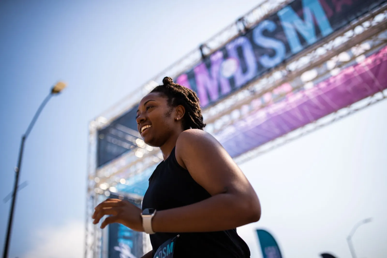 A smiling woman in a black tank top runs towards a race banner that says 'DSM'.