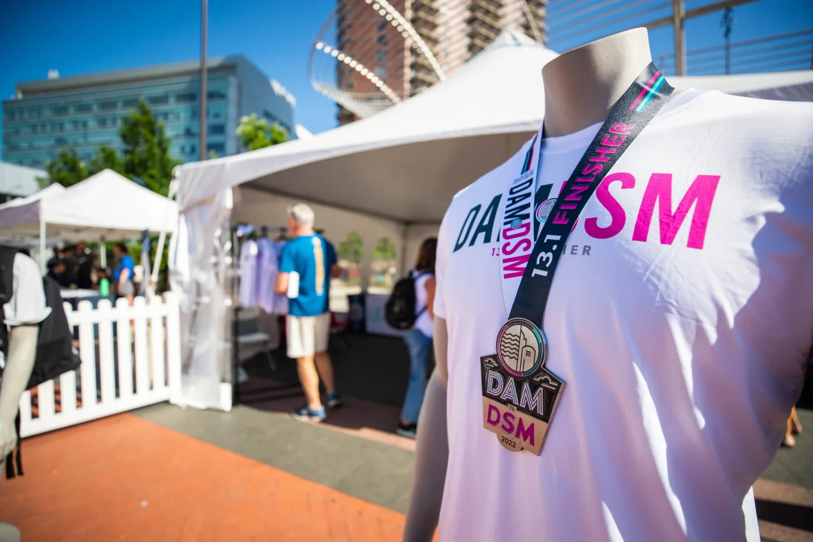 A mannequin wearing a white t-shirt with 'DAM to DSM' and a '13.1 FINISHER' medal around its neck, with tents and people in the background.