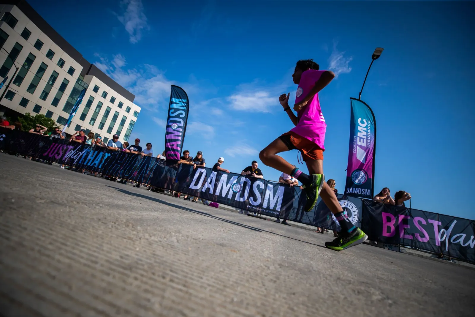 A runner in a pink shirt and orange shorts runs past spectators and race banners on a sunny day.