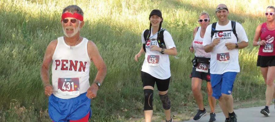A group of runners of various ages and genders participating in a race, with one runner in the foreground wearing a white tank top with 'PENN' on it and red and blue shorts.
