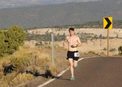 A male runner with bib number 56 runs shirtless on a paved road with a scenic desert landscape in the background.