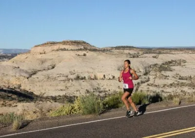 A female runner runs on a paved road with a scenic desert landscape in the background.