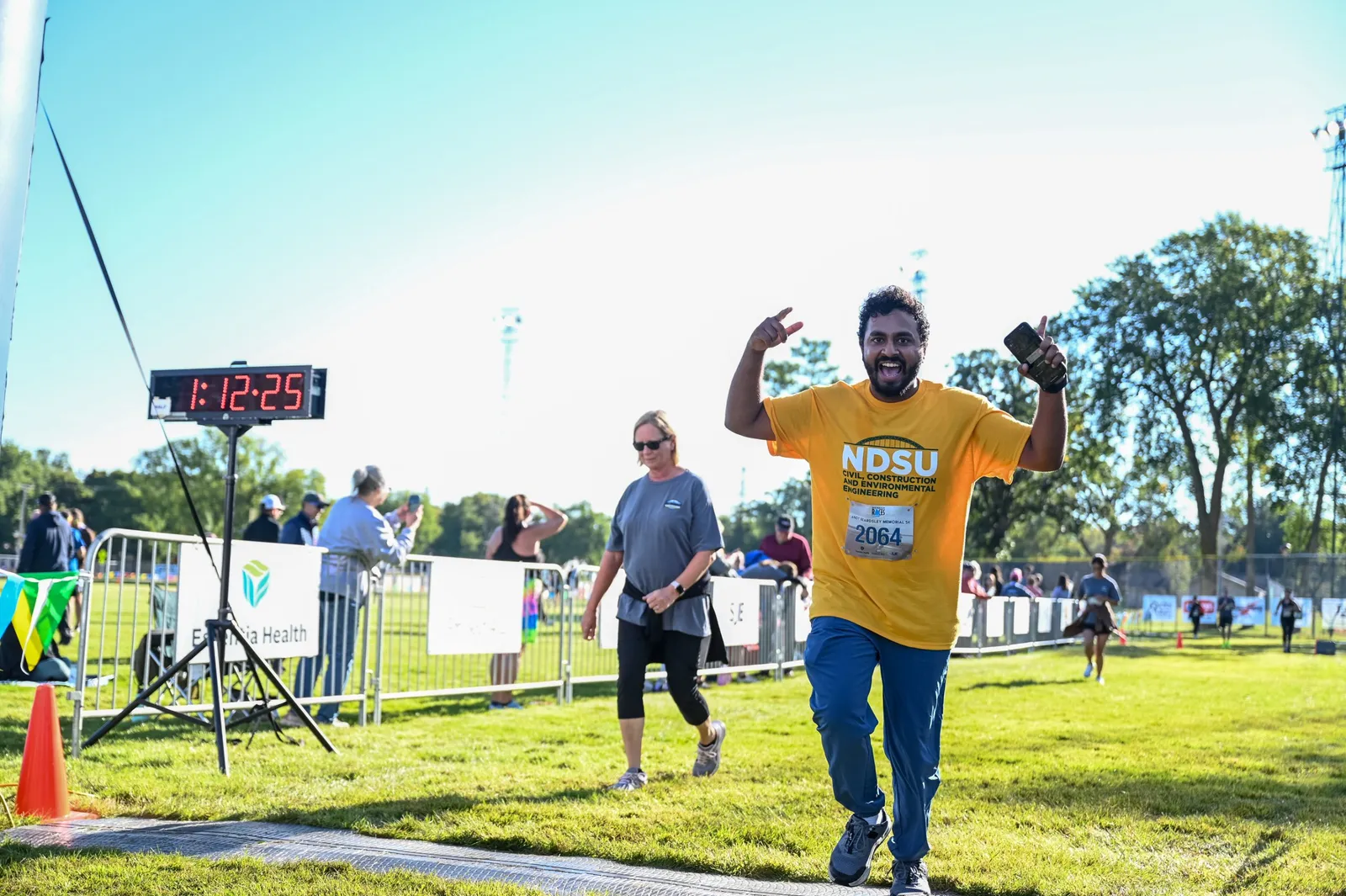 A runner in a yellow t-shirt crosses the finish line, celebrating with a peace sign and holding up a phone.