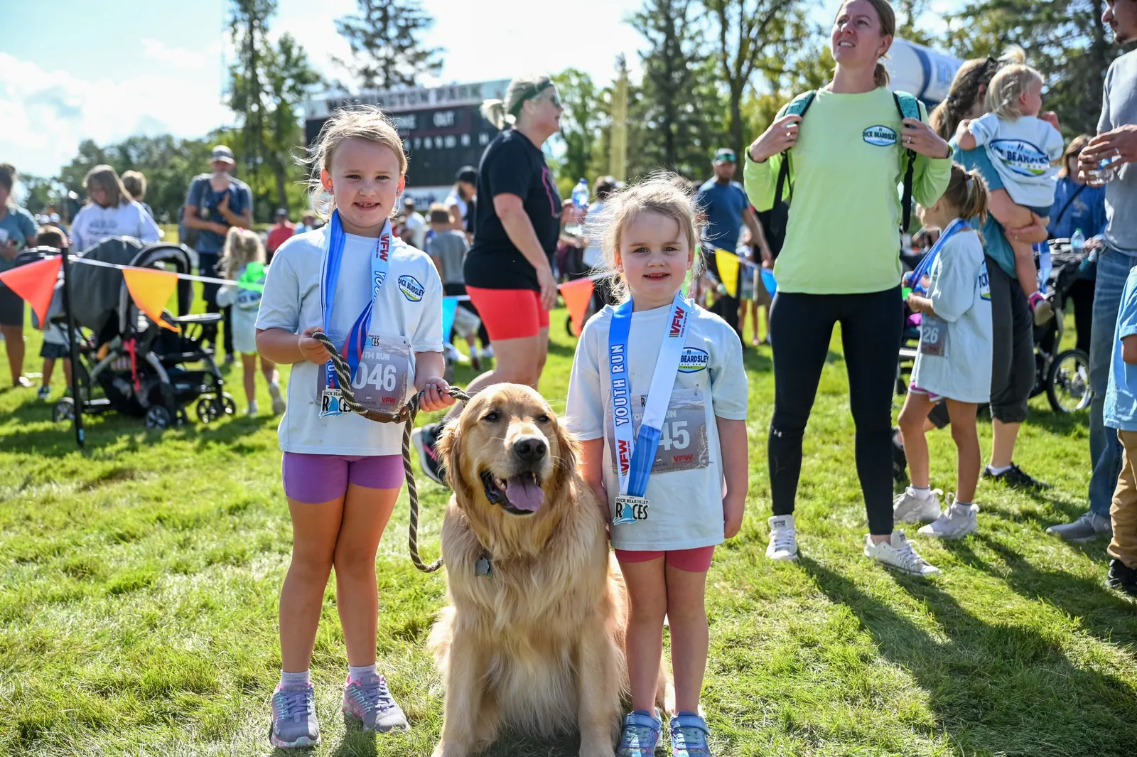 Two young girls with medals around their necks and a golden retriever dog posing for a photo at a race event.