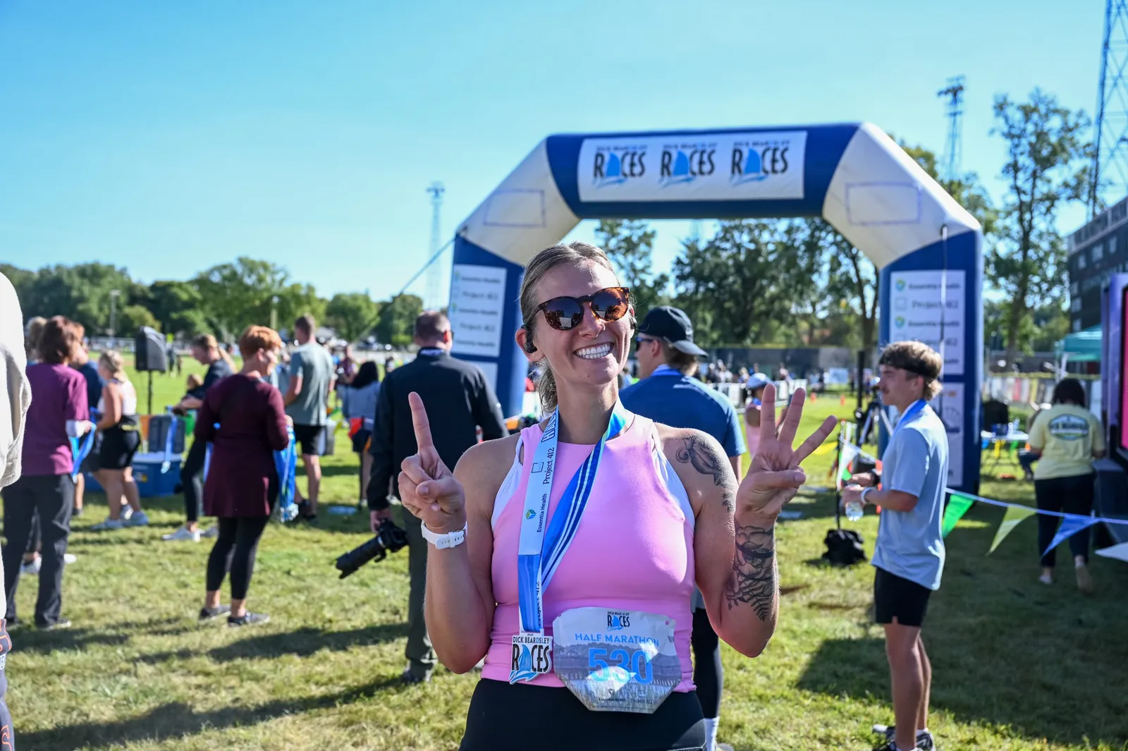 A smiling female runner wearing a pink tank top and sunglasses, with a half marathon medal around her neck, holds up two fingers in a peace sign and one finger up, standing in front of a blue inflatable race arch.