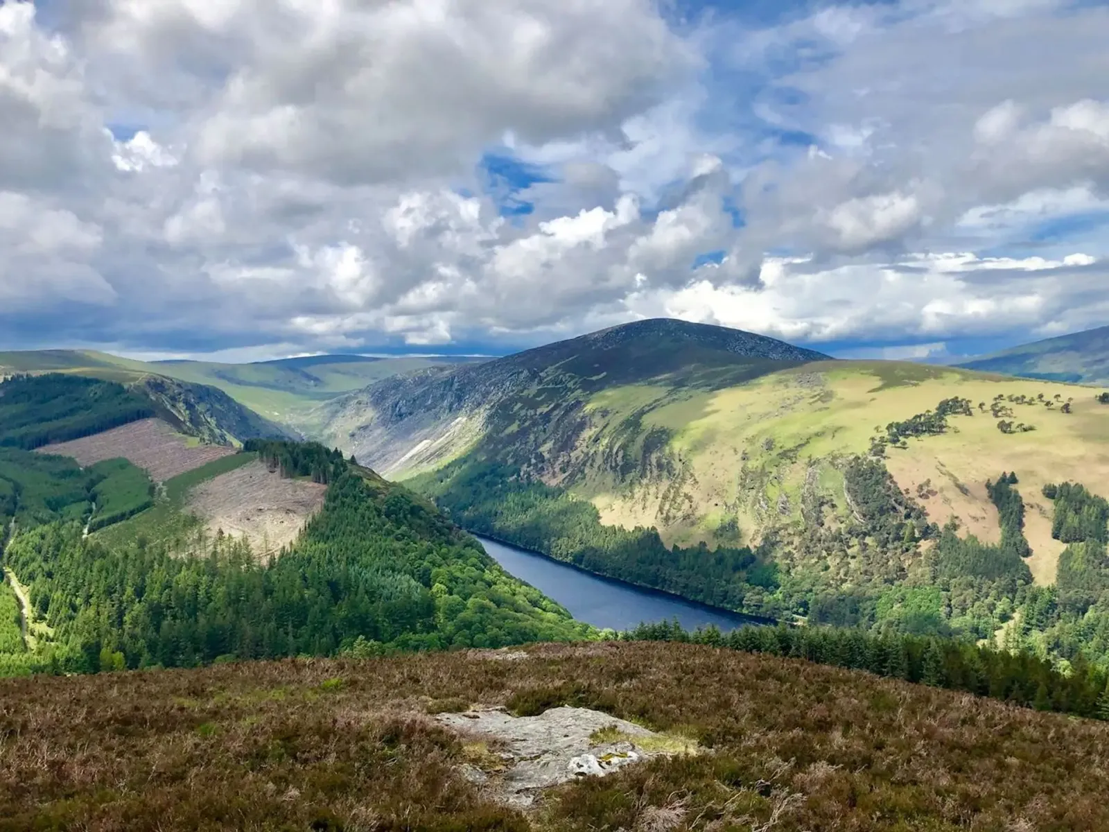 Scenic view of a valley with a lake, surrounded by mountains and forests under a cloudy sky.