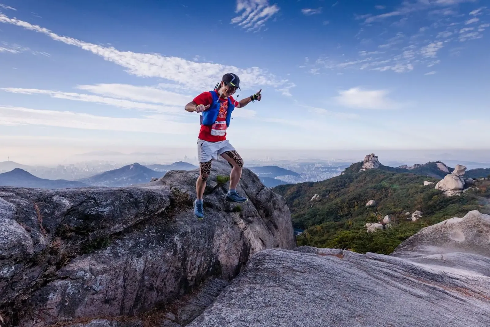 A trail runner gives a thumbs up while navigating a rocky mountain path with a city skyline in the distance under a blue sky with wispy clouds.