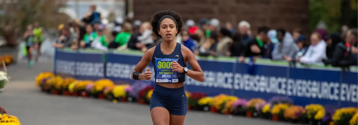 A female runner with bib number 3005 runs towards the camera, with spectators and race banners in the background.