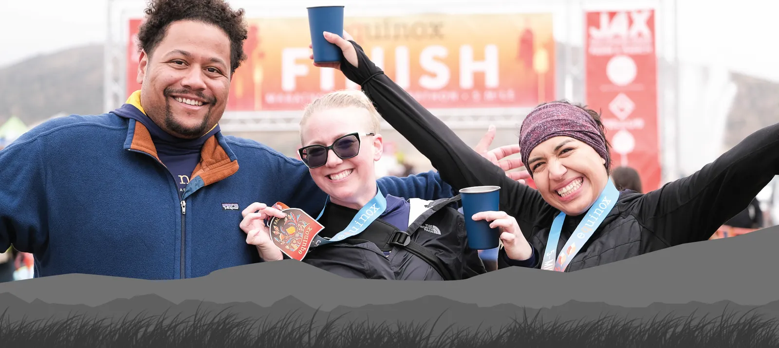 Three smiling runners, two women and one man, pose for a photo at the finish line of the Fall Equinox race, holding cups and wearing medals.