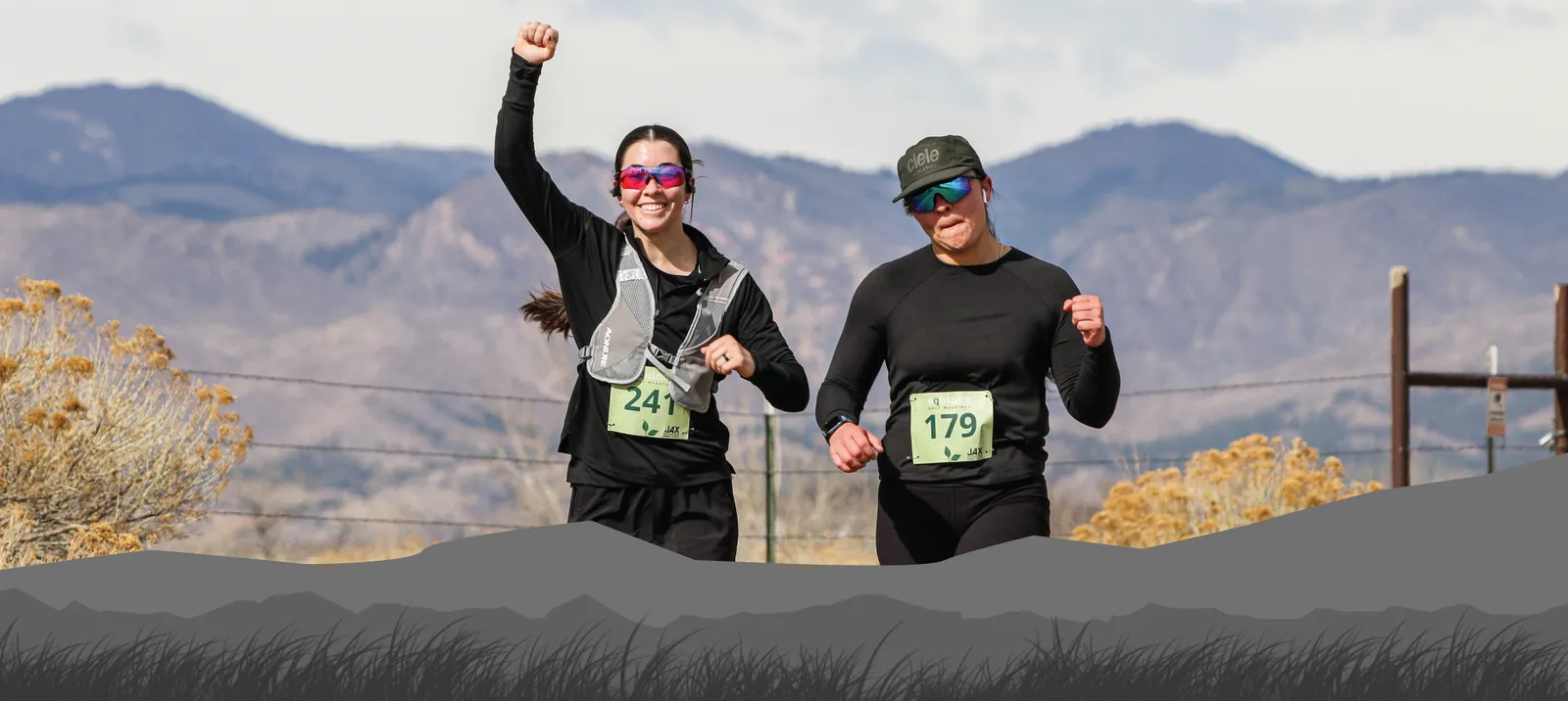 Two female runners are racing on a road with mountains in the background. The runner on the left is raising her fist in celebration.