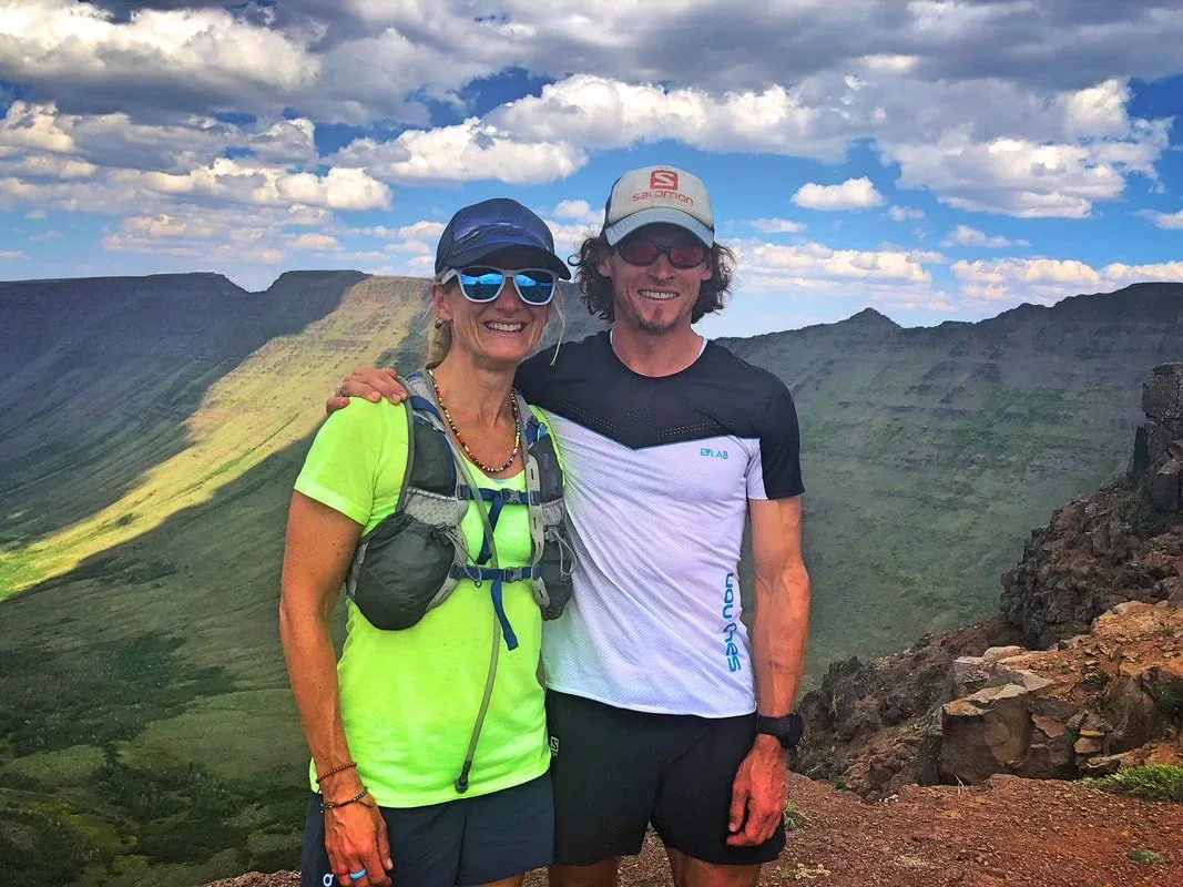 A man and a woman smiling at the camera, wearing running gear and standing on a mountain overlook with a scenic valley in the background.