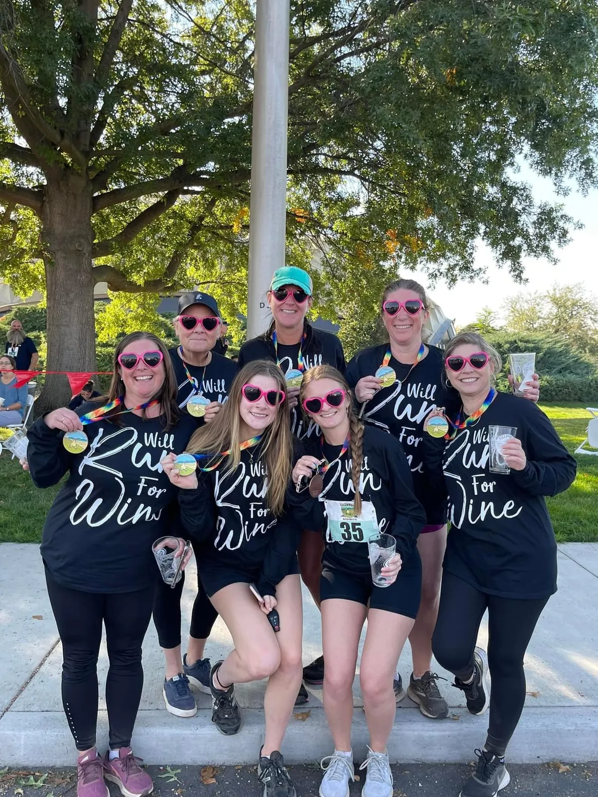 A group of seven women wearing black long-sleeved shirts and pink heart-shaped sunglasses, holding up their race medals and smiling.