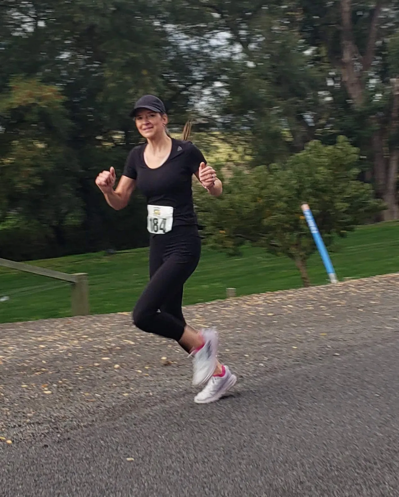 A female runner in black athletic wear and a cap runs on a paved path, with trees and grass in the background.