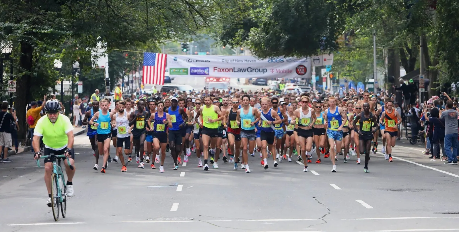 A large group of runners starting a road race, with a banner overhead and spectators on the side.