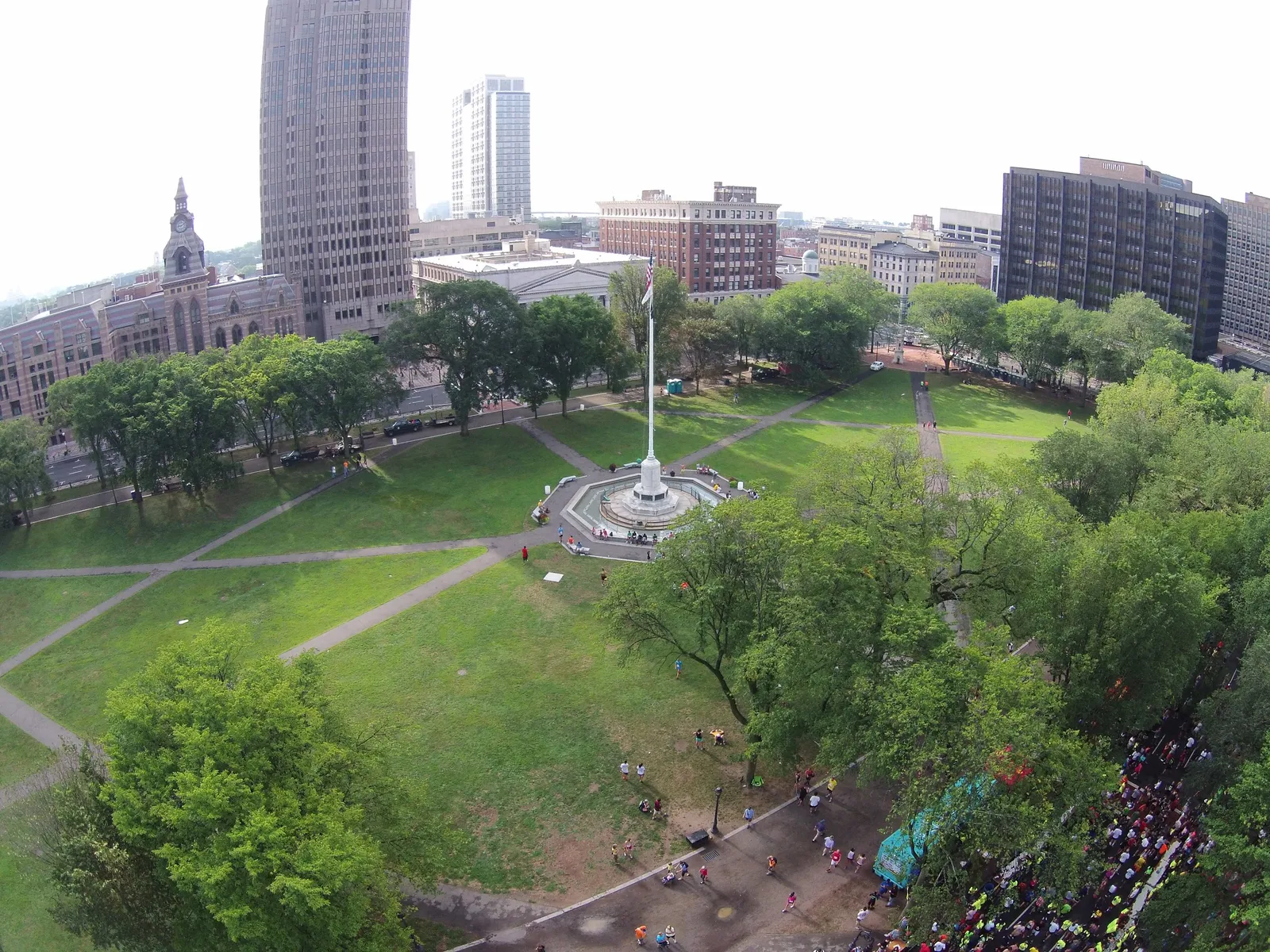 A crowd of runners are visible on a road next to a park with trees and buildings in the background.