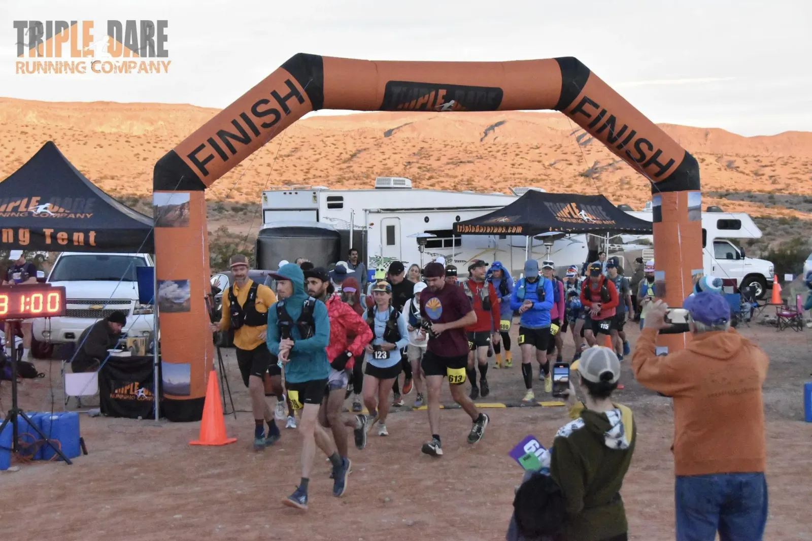 Runners starting a race under an inflatable arch with 'FINISH' and 'TRIPLE DARE' branding, with a desert landscape in the background.