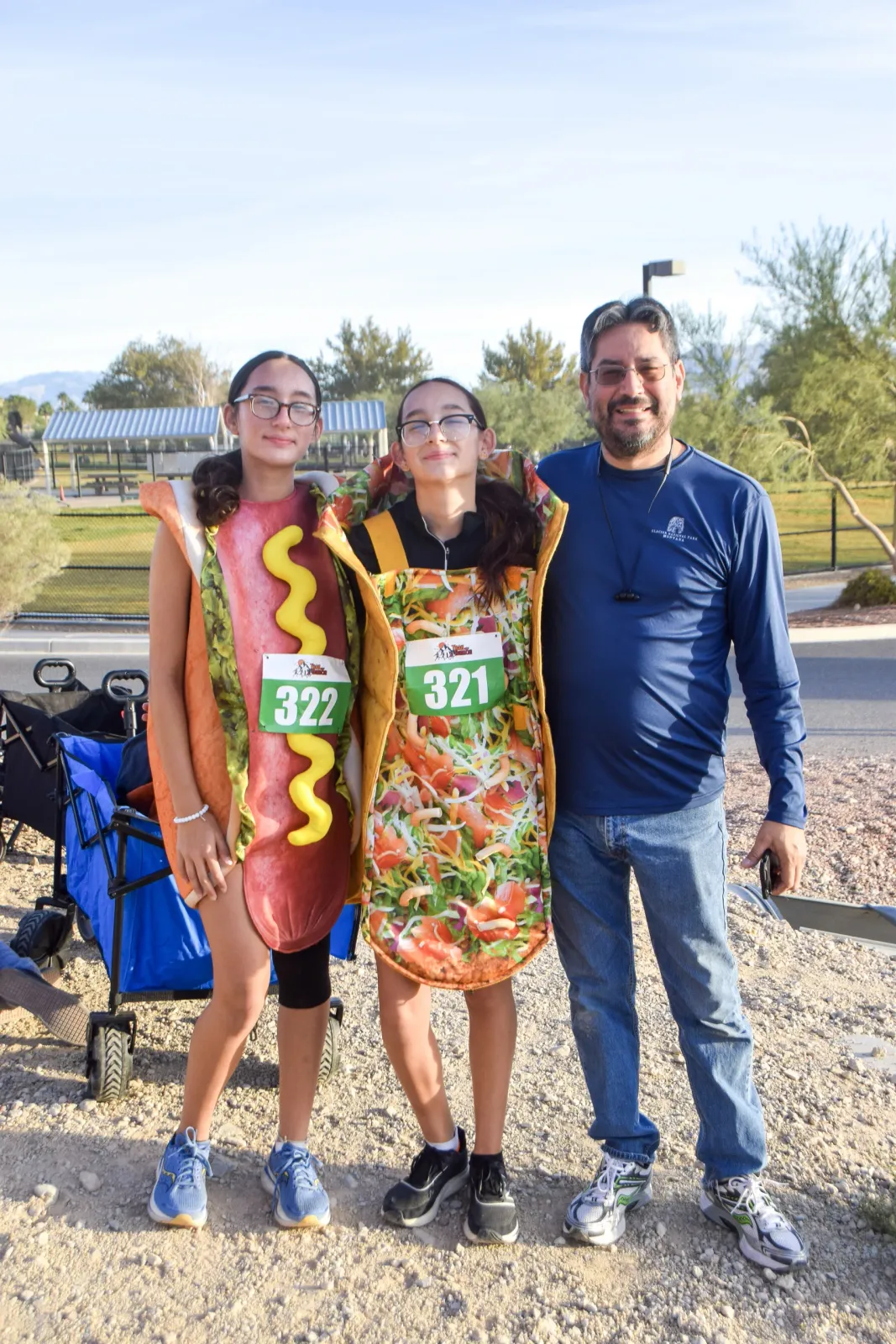A man and two young people in hot dog and taco costumes pose for a photo at a race, wearing bib numbers.