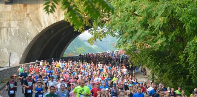 A large group of runners race along a paved path under a large stone bridge, with lush green trees lining the right side of the path.