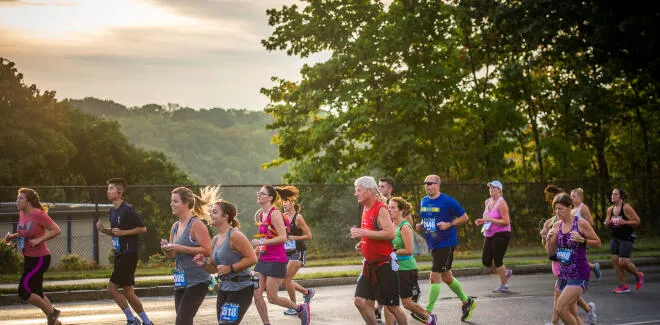 A group of runners of various ages and genders running on a road during a race, with trees and hills in the background under a cloudy sky.