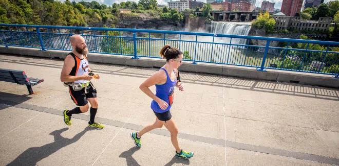 Two runners, a man and a woman, are running on a bridge with a waterfall and city skyline in the background.
