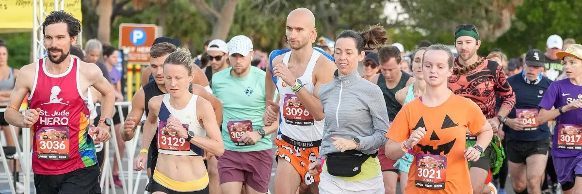 A group of runners, some in Halloween-themed costumes, race forward with bib numbers visible.