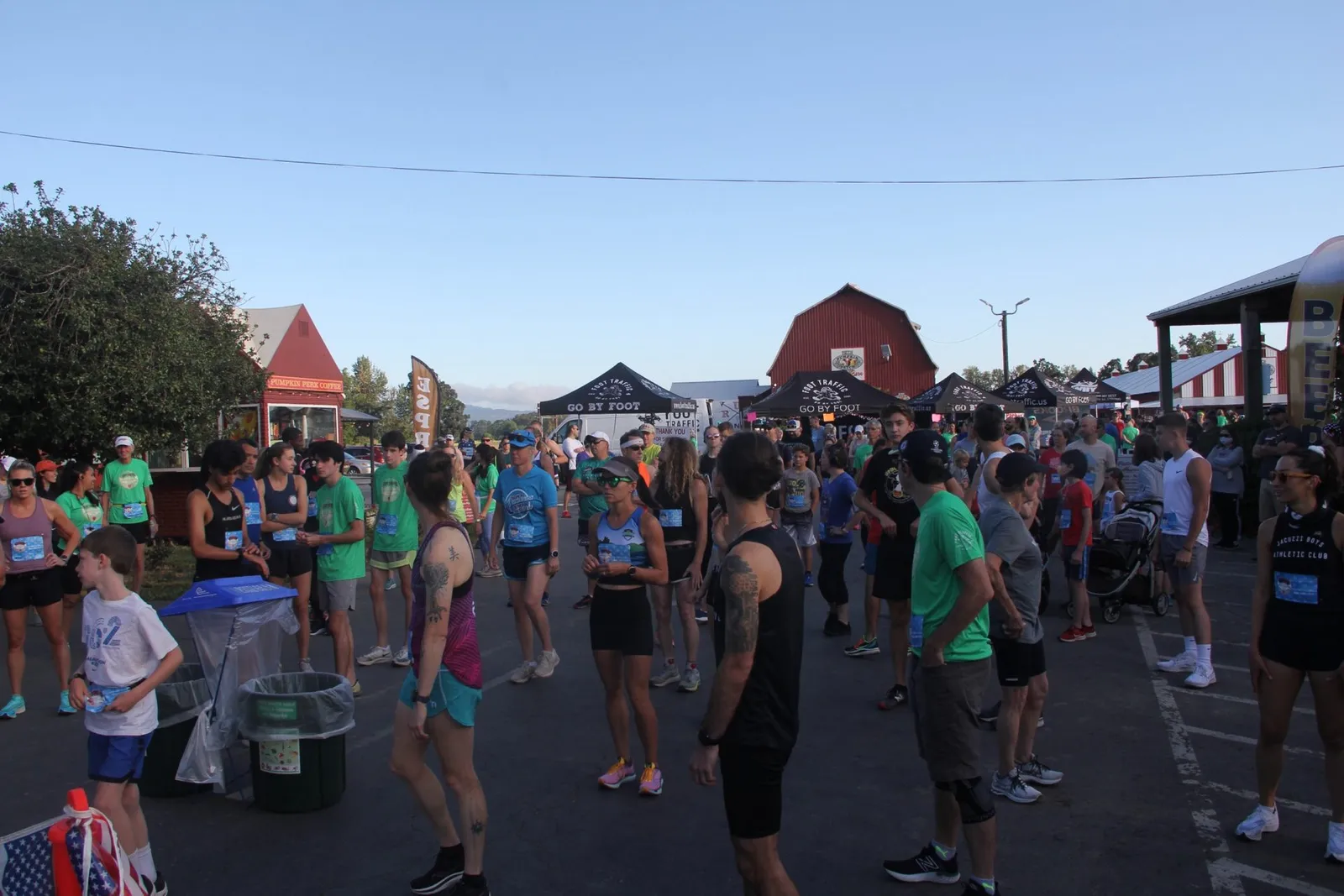A crowd of runners in athletic wear are gathered outdoors, with tents and a red barn in the background, appearing to be at the start of a race.