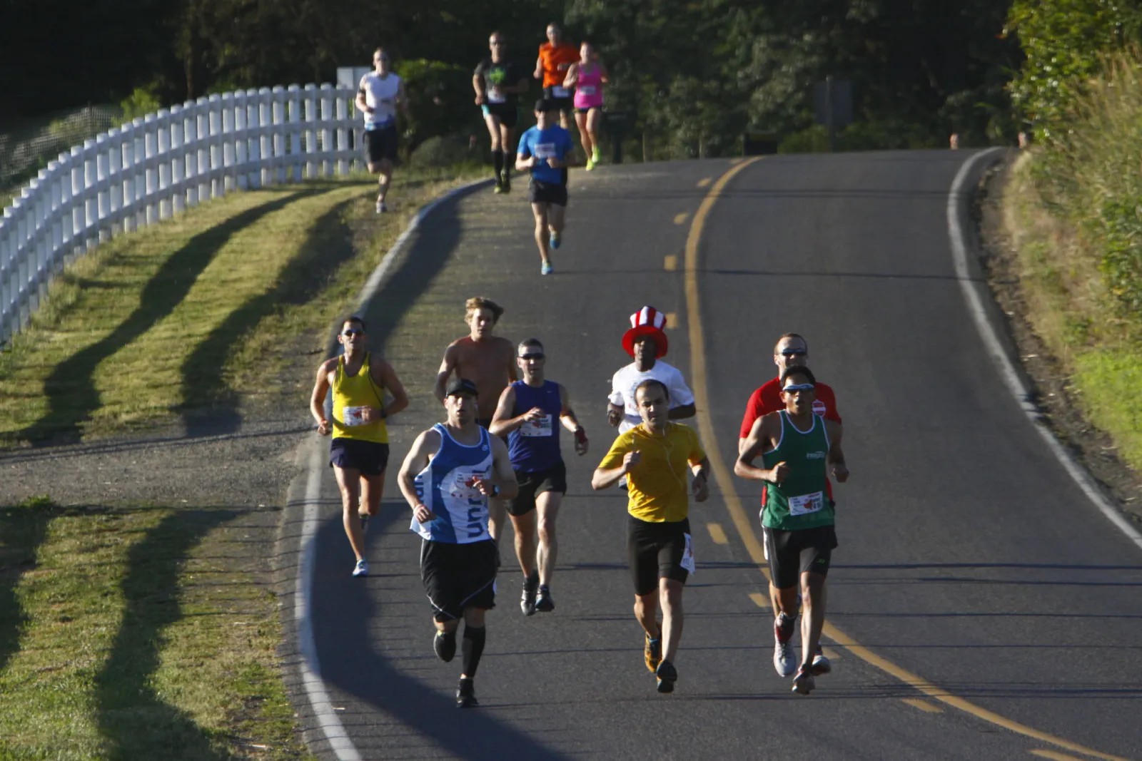 A group of runners race up a paved road with a white fence and grassy embankment on the left.